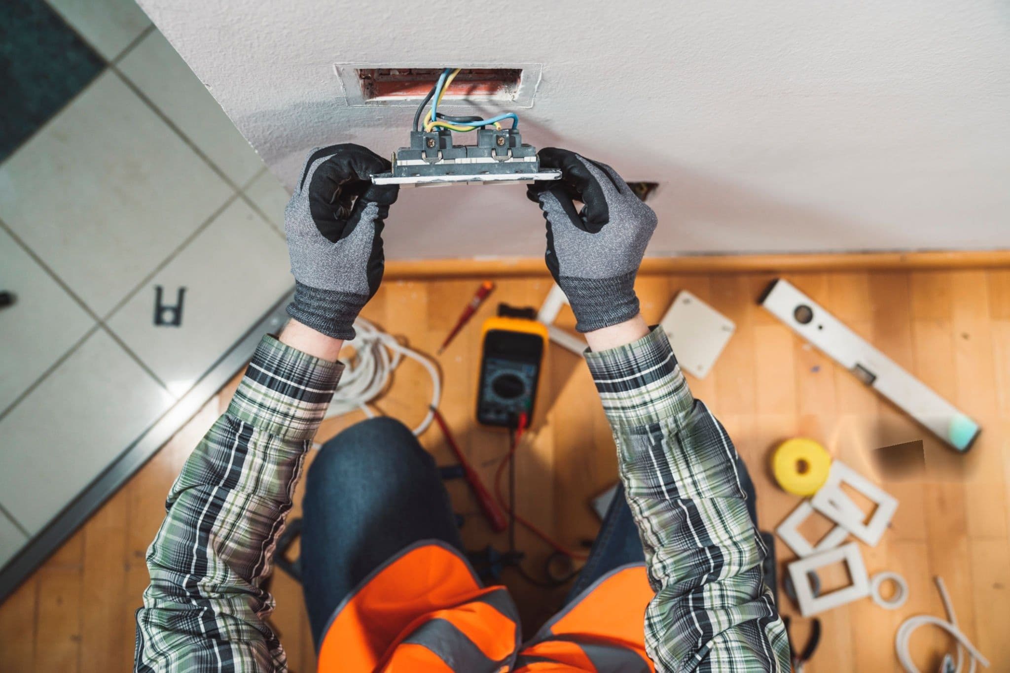 Overhead view of an electrician’s gloved hands fitting a wall socket, tools laid out on the floor below
